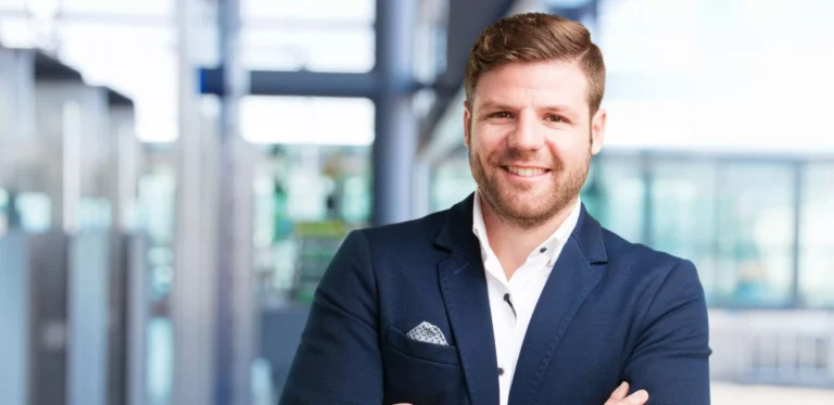 Homme souriant en costume, bras croisés, arrière-plan flou moderne.