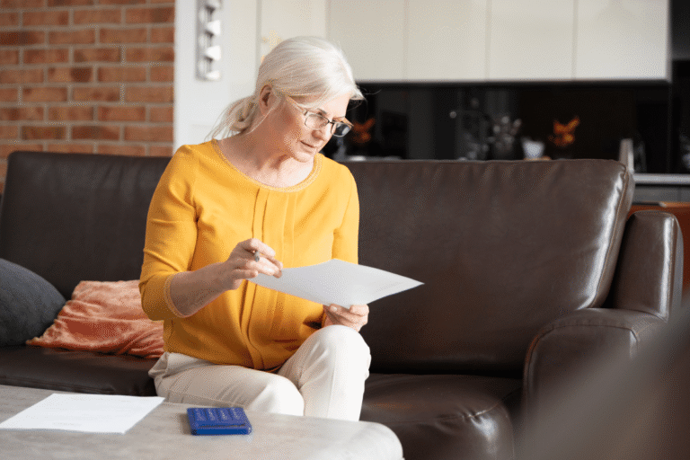 Femme âgée en pull jaune lisant un document assise sur un canapé.